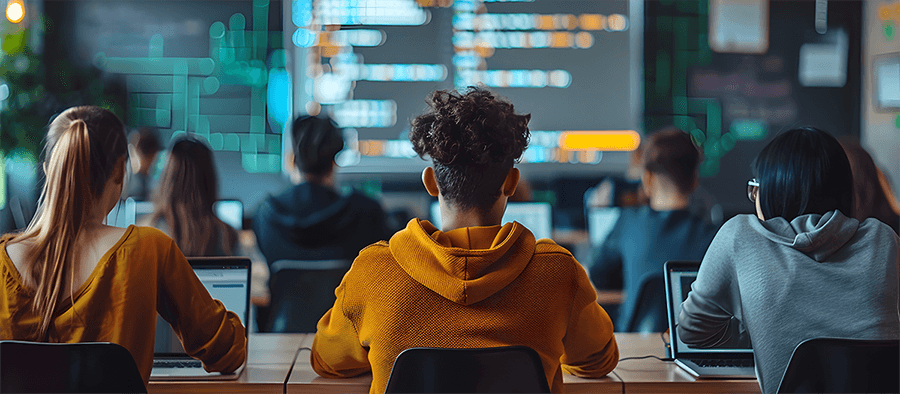 A class of students are all sitting in front of rows of laptops facing a white board which is displaying code