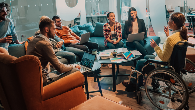 A group of office workers are sitting laughing together while working on laptops. A female wheelchair users is leading the meeting.