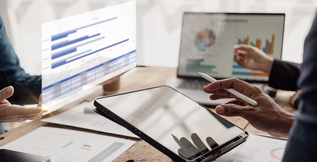 Three office workers are sitting around a desk assessing talent, equality, diversity, and inclusion data, which is displayed in graph and chart form, on varying devices