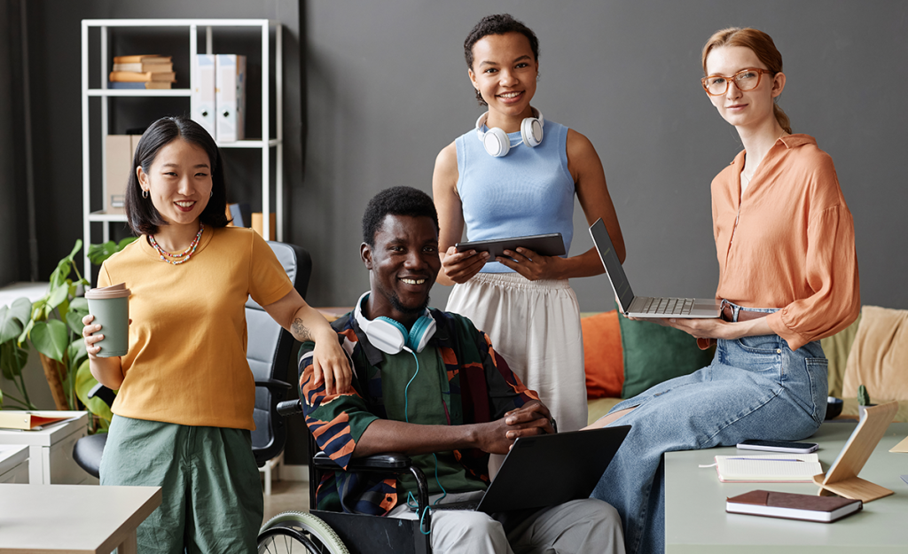 Four office workers, including a wheelchair users, are together all holding different digital devices such as tablets, laptops and headphones.