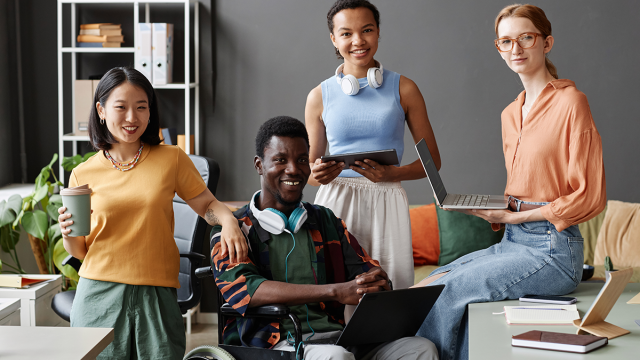 Four office workers, including a wheelchair users, are together all holding different digital devices such as tablets, laptops and headphones.